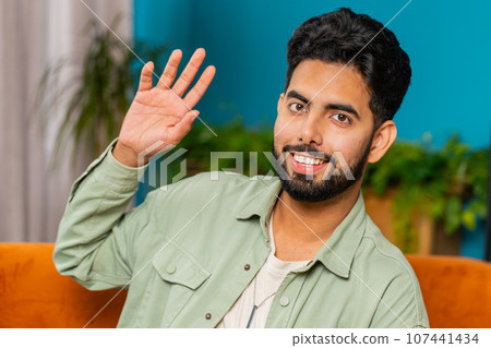 Indian man smiling friendly at camera, waving hands gesturing hello, hi, greeting at home on sofa 107441434