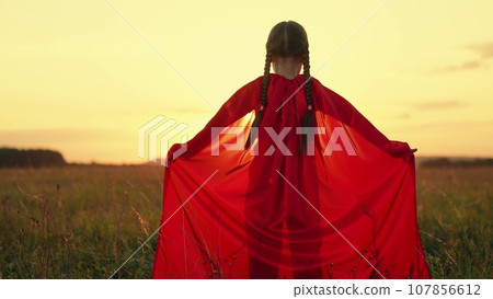 Cute little girl with braids wearing red cloak plays hero in sunset summer meadow 107856612