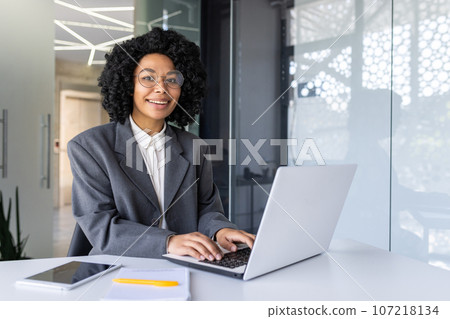 Portrait of young beautiful business woman in business suit, african american woman smiling and looking at camera, female worker with laptop sitting at table inside office. 107218134