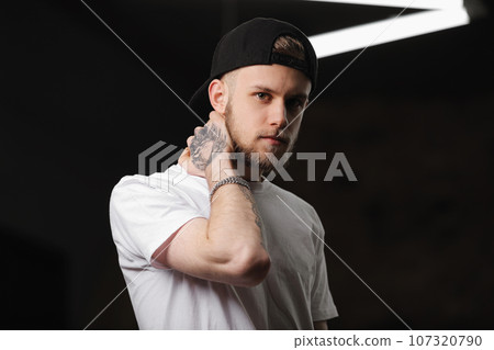 Stylish bearded caucasian young man with tattooed arm in cap and white t shirt looking at camera, light smiling, touching neck with hand on dark background with electric diode lamps. Fashion concept 107320790