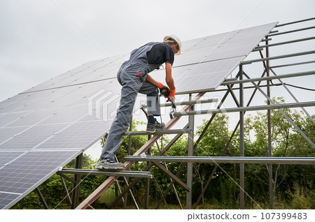 Worker fixing metal beams with electric drill for solar panels. Man wearing helmet and workwear. Photo-voltaic collection of modules as a PV panel. Array as a system of photo-voltaic panels. 107399483