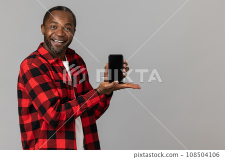 Joyful black bearded man wearing red checkered shirt showing smartphone with empty screen 108504106