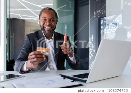 Mature successful businessman smiling and looking at camera, showing thumbs up, holding bank credit card, using laptop for online shopping and money transfer, inside office. 108374723