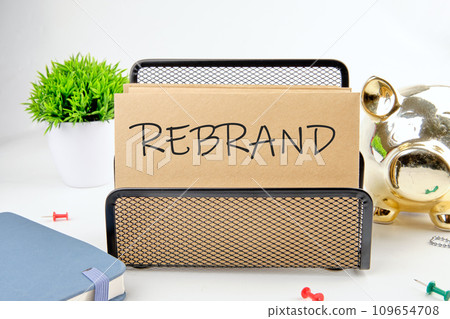 REBRAND text on paper next to piggy bank, notepad, flower on a white background 109654708