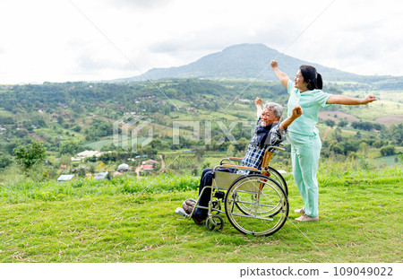 Side view of Asian nurse and senior man on wheelchair action of stretching together and both stay near the cliff with mountain and village as background. 109049022