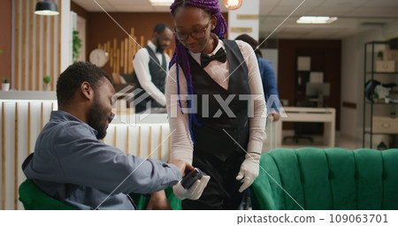 Traveller paying for coffee cup at hotel, using card at pos terminal to buy refreshment from bar. Client thanking employee for serving beverage in lounge area while he waits for check in. 109063701