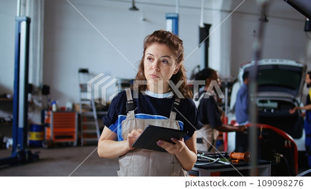 Cheerful engineer in repair shop using tablet to check car performance parameters during inspection. Relaxed garage workspace employee using device to examine damaged vehicle 109098276