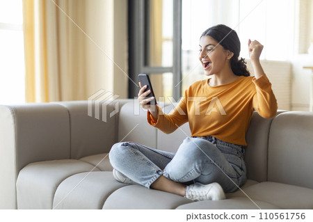 Overjoyed indian woman with smartphone in hand celebrating success at home 110561756