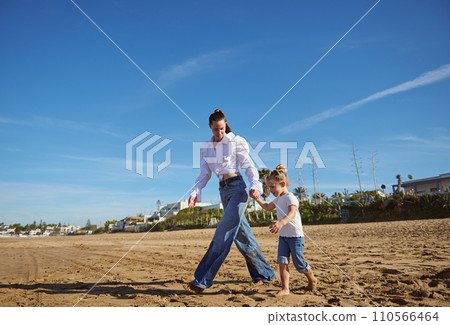 Young loving mother and daughter holding hand, walking barefoot together on the sandy beach. People. Leisure activity 110566464