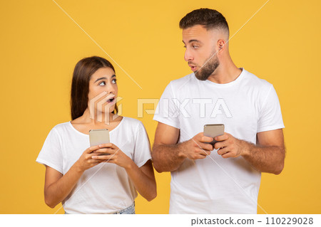 Young woman and man in white t-shirts, both holding smartphones with surprised expressions 110229028