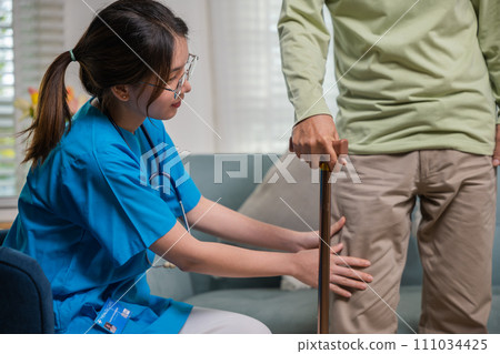 Asian young woman nurse checking knee and leg after surgery of senior old man patient suffering from pain in knee, doctor asking elderly man about pain symptom with walking stick 111034425