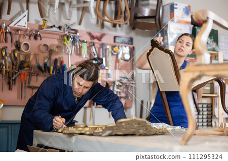 Male restorer treats wooden frame of vintage mirror with protective varnish. While woman restorer restores upholstery of vintage chair 111295324