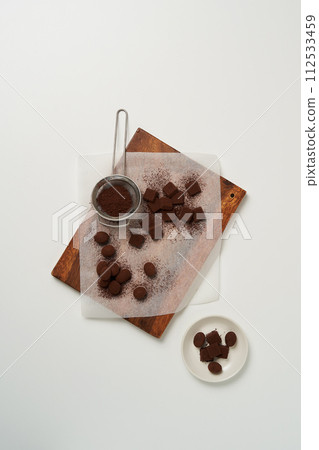 There are pieces of homemade chocolate sprinkled with cocoa powder on the cutting board and a strainer on the side 112533459