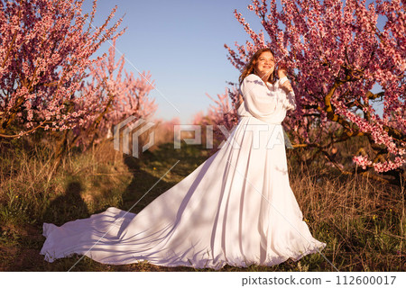 Woman blooming peach orchard. Against the backdrop of a picturesque peach orchard, a woman in a long white dress enjoys a peaceful walk in the park, surrounded by the beauty of nature. 112600017