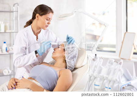 Aesthetician administering facial procedure on young woman using ultrasonic shovel 112602801