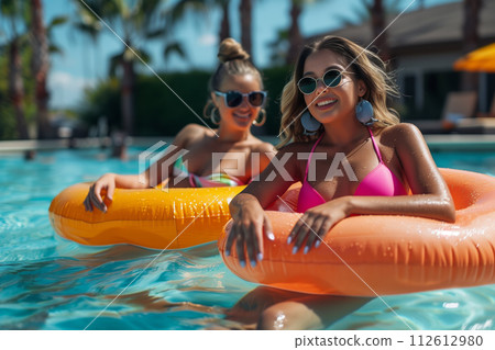 Two happy women in sunglasses lounging on pool floats under the sun 112612980