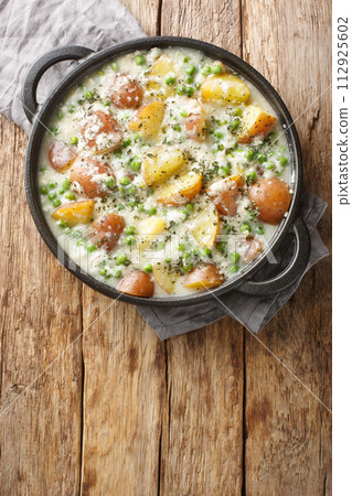 Vegetarian baked potatoes and green peas in creamy cheese sauce close-up in a pan on a wooden table. Vertical top view 112925602