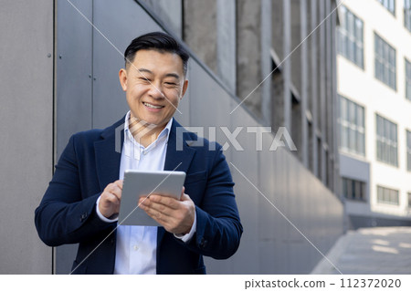 Smiling Asian businessman in a blue suit using a digital tablet outside an office building on a sunny day. 112372020