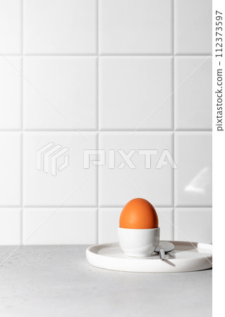 Boiled egg in an egg stand and spoon on white tile kitchen background with text space. Light protein breakfast concept, minimalism, high key 112373597