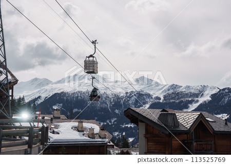 Winter wonderland scene at a ski resort with wooden chalet buildings, ski lift, snow covered mountains, and ski runs. Serene and chilly atmosphere. Located in European Alps or similar region. 113525769