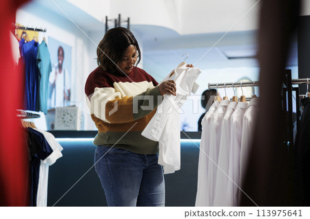 Fashion boutique client holding white shirt on hanger and checking fabric quality. African american woman examining apparel rack while searching for formal outfit in clothing store 113975641