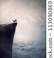 Superhero with cape stands brave on edge of a cliff above the clouds searching determined at horizon with hand up to forehead. Powerful hero conceptual scene 113090869