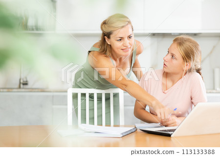 Mother looks at laptop and prompts and helps teen girl to write in notebook to complete task 113133388