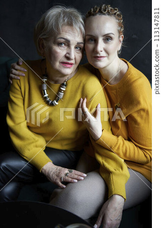 stylish old mother and middle-aged daughter in yellow sweaters hug and comfort each other in a cafe 113147811