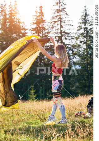 Woman traveller camping outdoors. Female tourist wearing sportswear, putting tent, making campsite in mountains. Concept of tourism and beauty of nature. 113238058