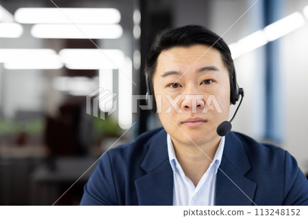 Close-up portrait of a serious young Asian man wearing a headset sitting in the office and looking confidently into the camera. 113248152