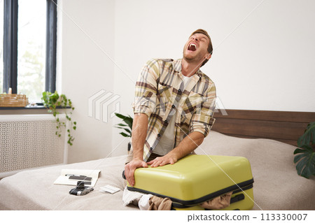 Portrait of man struggling to close suitcase full of clothes, putting effort to pack luggage for holiday, going on long vacation with lots of things 113330007