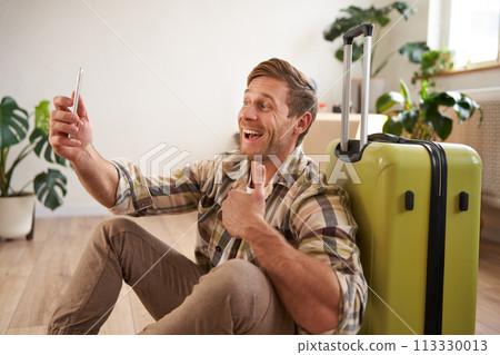 Portrait of happy tourist, man takes selfies before his flight, photos with suitcase, uses smartphone to post pictures on social media 113330013