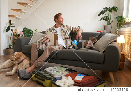 Portrait of young man with check list of items in hands, goes on holiday with his dog, packs suitcase for vacation 113333138