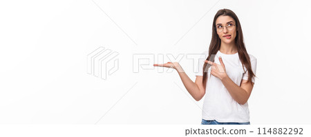 Waist-up portrait of thoughtful, pretty young woman holding arm as if have something lying in it, pointing at product with pondering face, bite lip look up thinking, white background 114882292