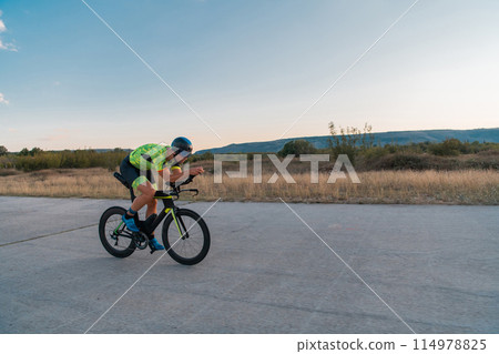 Triathlete riding his bicycle during sunset, preparing for a marathon. The warm colors of the sky provide a beautiful backdrop for his determined and focused effort. 114978825