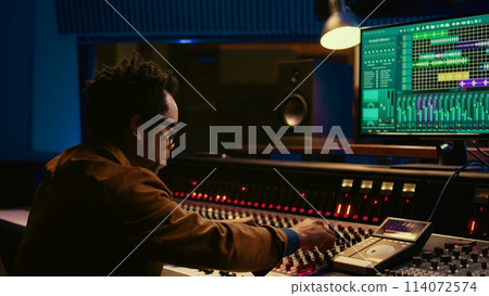 Sound designer uses mixing console and managing audio tracks, twisting knobs on control desk for adjusting tunes and volume. Producer creates music in professional studio, technical gear. Camera B. 114072574