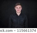 Portrait of a carefree young man against a black wall background looking to camera. Relaxed male model with joyful face expression wearing dark blue jeans jacket 115661374