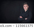 Portrait of a confident young man, keeps arms folded against a black wall background with copy space. Successful male student with determined face expression wearing dark blue jeans jacket 115661375