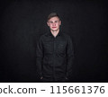 Confident and relaxed young man portrait, keeps hands in pockets against a black wall background. Carefree male student with positive face expression wearing dark blue jeans jacket 115661376