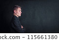 Pensive young man side view portrait, keeps arms folded against a dark wall background with copy space. Thoughtful male student with determined face expression 115661380