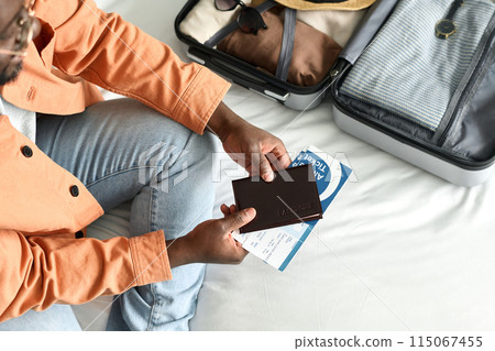 Top view close up of unrecognizable Black man holding passport and tickets sitting on bed in hotel room and preparing for travel copy space 115067455