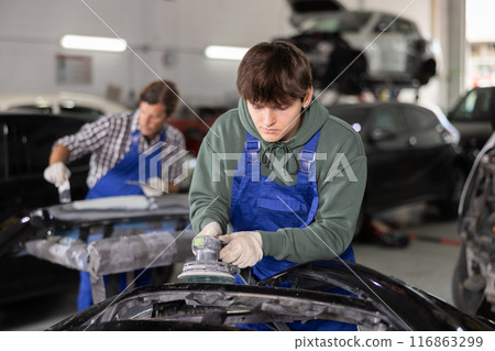 Young auto technician sanding car part with power sander in workshop 116863299