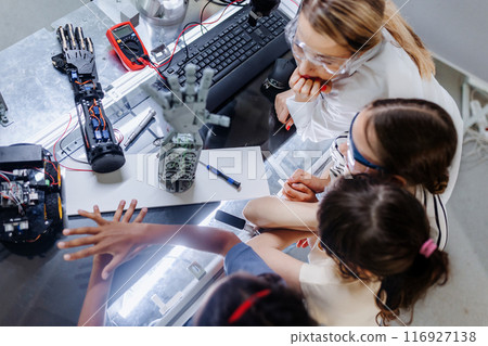 High angle view of teacher helping to girls working on small robot, building robotic kit in after-school robotics club. Children learning robotics in Elementary school. Girls in science. 116927138