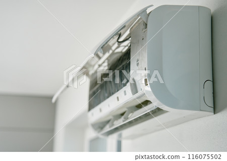 White air conditioner unit is mounted on wall, ready for maintenance. Climate system with opened front cover revealing internal components. Repairing HVAC in apartment 116075502