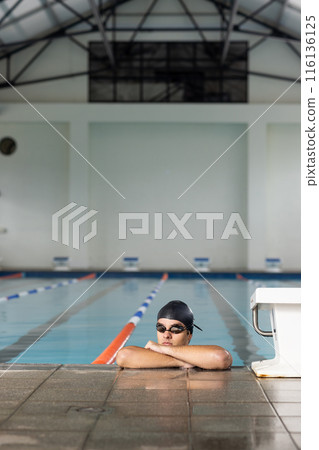 Caucasian young male swimmer resting at pool edge indoors, looking away, copy space 116136125