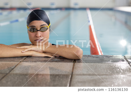 Young biracial female swimmer resting at pool edge indoors, wearing goggles, copy space 116136130