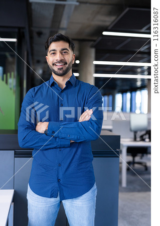 Smiling businessman in modern office, standing with arms crossed, wearing blue shirt 116316087