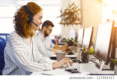 A woman wearing a headset is seated in front of a computer, appearing focused on her work. She is typing on the keyboard and looking at the screen intently 117495317