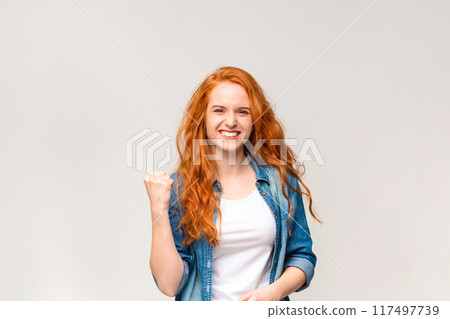 A radiant young woman with flowing red hair is captured mid-celebration, her fist pumped in victory and a beaming smile across her face, signaling triumph or good news 117497739