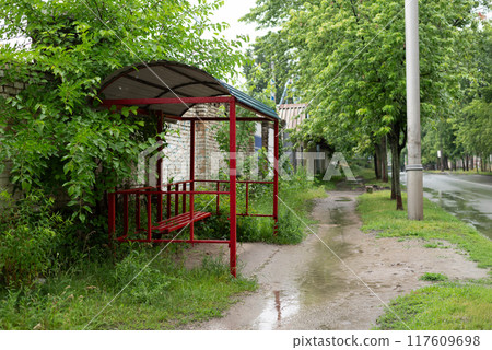 Bus stop on the side in the countryside, or rural area. Bus stop is empty. Rainy day. There Is green grass and trees. 117609698